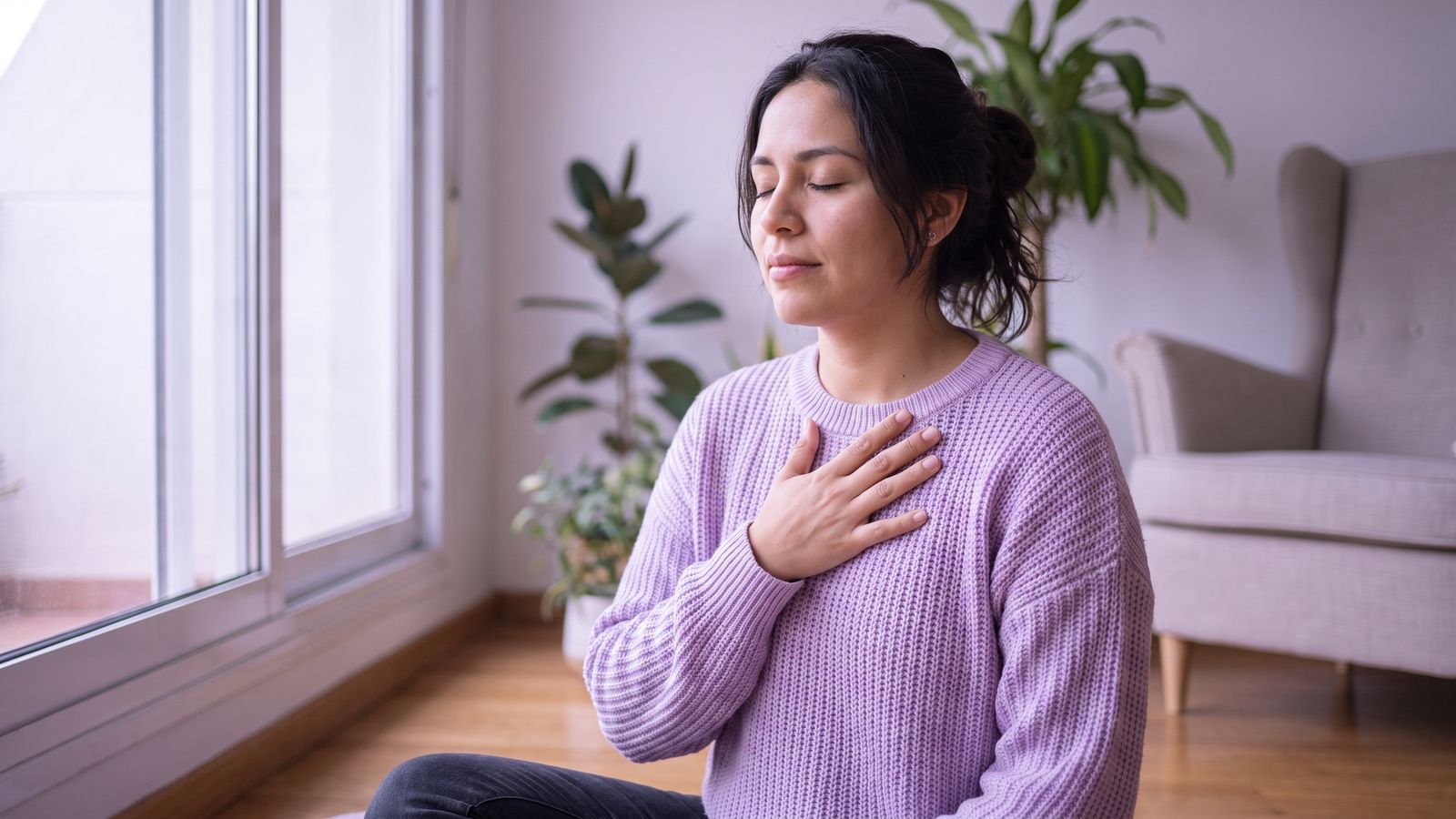 Mujer sentada junto a la ventana respirando profundo con la mano en el pecho